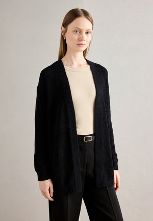Cardigan en tricot noir avec une ouverture sur le devant, des manches longues et une surface texturée ; porté sur un haut beige et un pantalon noir.