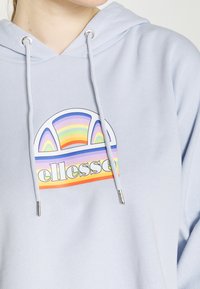 Felpa con cappuccio azzurro chiaro, con una grafica frontale raffigurante un arco di colore arcobaleno e la parola "ellesse" in caratteri in grassetto. Aggiuni tiranti con punte in argento.