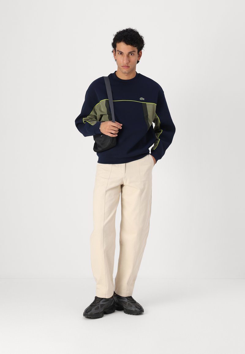 Sweatshirt bleu marine avec des accents verts, pantalon crème et baskets noires. Le modèle porte un petit sac noir, se tenant sur fond blanc.