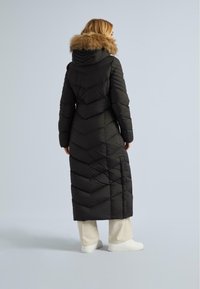 O femeie îmbrăcată cu un palton lung negru, matlasat, cu glugă căptușită cu blană, pantaloni crem și adidași albi, stând pe un fundal simplu.
