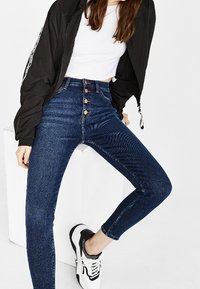 Vaqueros de tiro alto en azul con un corte ajustado, que presentan un cierre delantero de cinco botones y una sutil textura de espiga. Combinados con una chaqueta bomber negra.
