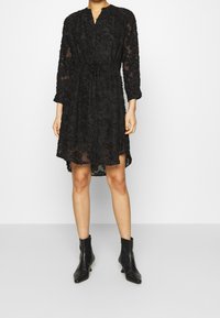 Vestido de encaje negro con mangas largas, cinturón en la cintura, textura floral y un diseño ligeramente transparente, combinado con botines negros. 