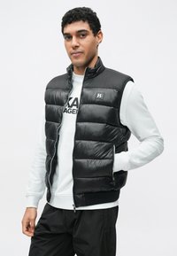 Čierna prešívaná vesta s vodorovným prešívaným vzorom, vysokým golierom a zapínaním na zips. Nosená cez bielu mikinu s bočnými vreckami.