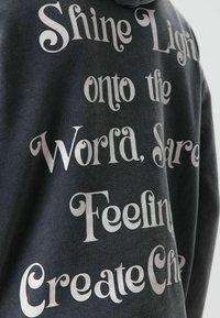 Sweat à capuche gris foncé avec un texte argenté audacieux dans une police ludique. Le texte dit : « Illumine le monde, Partage des émotions, Crée de la joie. »