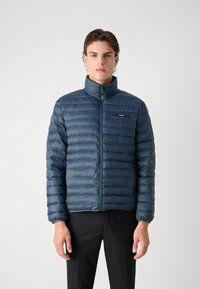 Giacca puffer blu con design a righe orizzontali, colletto alto e chiusura a zip sul davanti. Presenta un patch con logo sul lato sinistro del petto. Indossata sopra pantaloni neri.