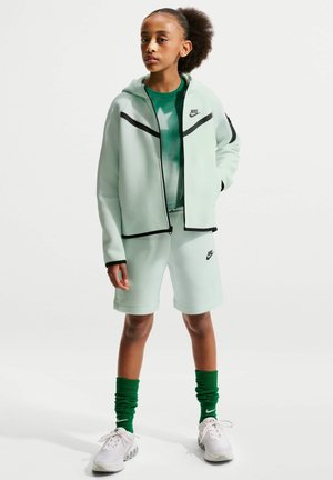 Ung mand iført lysegrøn Nike hættetrøje, matchende shorts, grøn stjernemønstret skjorte, grønne høje sokker og hvide sneakers, stående med hånden i lommen.