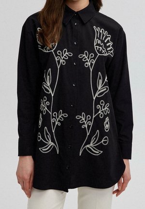 Femme portant une chemise noire boutonnée avec une broderie florale blanche sur le devant, associée à un pantalon clair.
