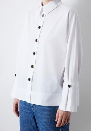 Valkoinen pitkähihainen paita, jossa mustat ja kultaiset neliönmuotoiset napit, käytetään paita housujen ulkopuolella sinisten farkkujen kanssa, kädet rentoina sivuilla.