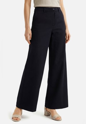 Femme portant un pantalon noir à jambes larges associé à des sandales beige à talon bloc ouvertes et un haut beige, debout devant un fond uni.