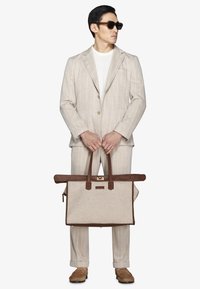 Uomo con completo beige a righe e camicia bianca che indossa occhiali da sole, tiene una grande borsa tote in pelle beige e marrone, in piedi su sfondo bianco.