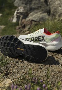 Ζευγάρι λευκά και πορτοκαλί παπούτσια trail running Terrex σε βραχώδες έδαφος, το ένα δείχνει μαύρη ελαστική σόλα Continental με βαθιές εμπρόσθιες εγκοπές.