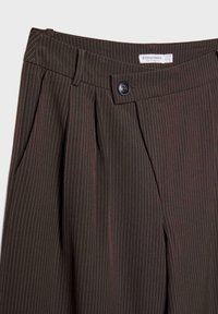 Pantalon marron foncé à fines rayures avec plis, poches latérales, fermeture par bouton à la taille et étiquette de marque visible à l'intérieur de la ceinture.