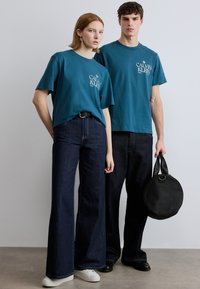 Βαμβακερά μπλουζάκια τιρκουάζ με λευκό λογότυπο, σε συνδυασμό με σκούρο τζιν με φαρδιές παντελόνες. Ένα μοντέλο κρατά μια στρογγυλή μαύρη τσάντα, και οι δύο φορούν casual παπούτσια.