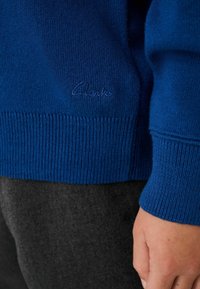Blauwe gebreide trui met ribgebreide manchetten en zoom. Bevat een subtiele reliëf "Clarks" logo. Grijze stof zichtbaar eronder. Zachte textuur.