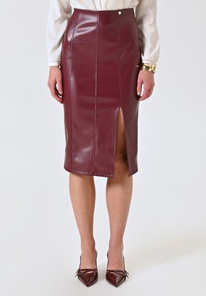 Femme portant une jupe crayon en cuir bordeaux avec une fente frontale, associée à des talons pointus assortis, un chemisier blanc et un bracelet en or.