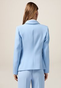 Blazer su misura azzurro chiaro con colletto a scollo, silhouette aderente e texture liscia, dotato di una fessura sul retro per facilitare i movimenti.