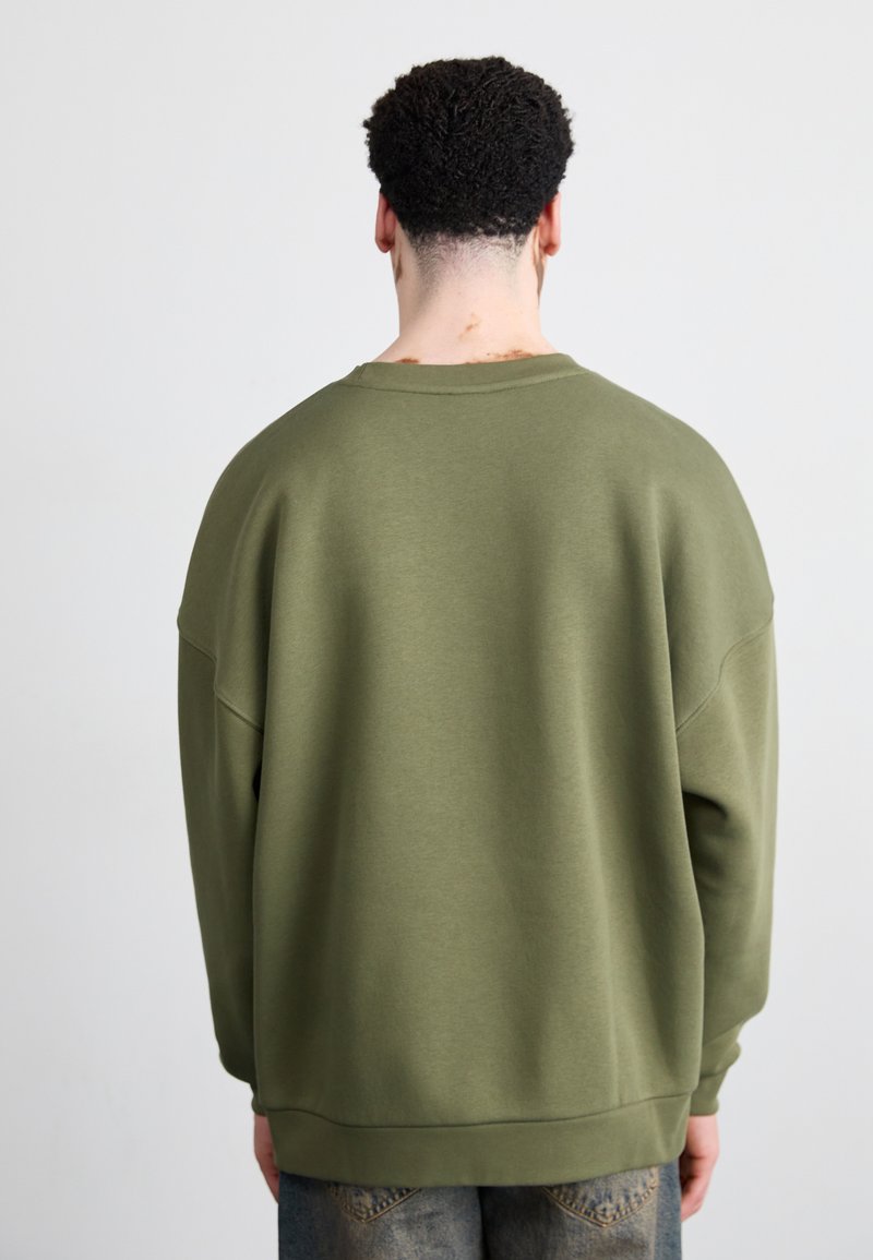 Felpa verde oliva con vestibilità ampia, spalle ribassate e polsini a coste. Texture liscia e design semplice, senza motivi o accenti evidenti.