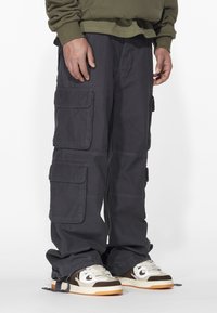 Cargo pants in dark gray fabric with multiple pockets, loose fit, and ankle cuffs. Paired with sneakers featuring beige and brown accents.
