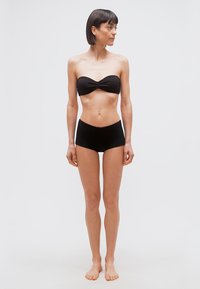 Γυναίκα με κοντά σκούρα μαλλιά στέκεται ξυπόλυτη, φοράει μαύρο στράπλες μπαντό τοπ και μαύρο σορτς ψηλόμεσο, μπροστά σε λευκό φόντο.