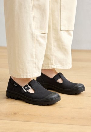 Fekete vászoncipők, kivágott mintázattal, gumitorlóval és állítható csattal. Texturált talp barázdált mintázattal. Világos nadrággal viselve.