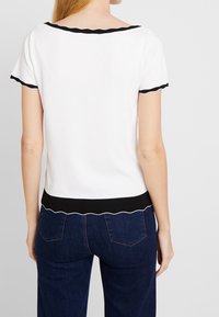 Mujer con una blusa blanca de manga corta con bordes negros en forma de concha y jeans de color azul oscuro, vista desde atrás, de pie contra un fondo liso.