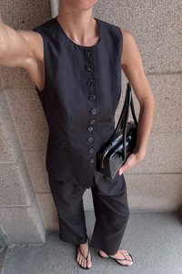 Zwarte mouwloze blouse met knopen aan de voorkant, gecombineerd met losse zwarte broek en een kleine zwarte handtas. Sandalen maken de outfit compleet.