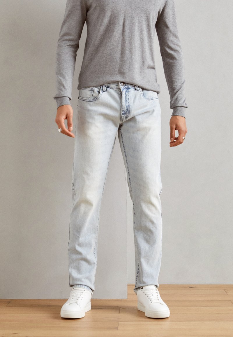 Light wash denim jeans with a straight leg fit, front pockets, and visible stitching, paired with a gray long-sleeve shirt and white sneakers.