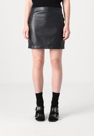 Jupe mini en cuir noir avec une texture lisse et une coupe ajustée. Associée à des bottines noires avec des boucles dorées et des chaussettes noires.