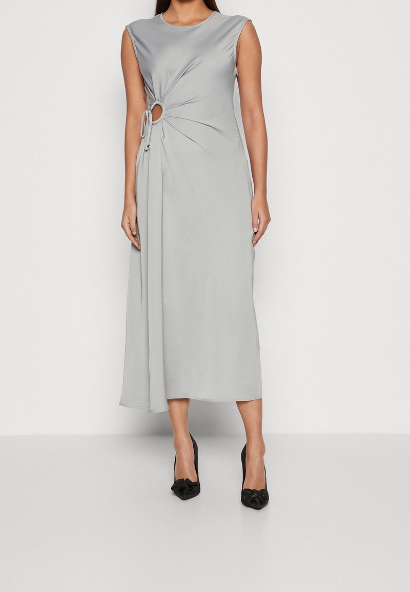 Femme portant une robe midi sans manches gris clair avec une découpe latérale et des fronces, associée à des escarpins noirs à bout pointu avec des nœuds.