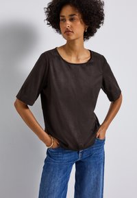 Donna con capelli ricci che indossa un top scuro a maniche corte e jeans blu, in piedi con le mani nelle tasche di fronte a un muro bianco uniforme.