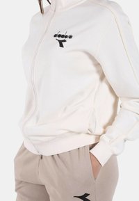 Giacca bianca con zip e logo nero, abbinata a pantaloni della tuta beige. Tessuto morbido, vestibilità standard, con tasche visibili sia sulla giacca che sui pantaloni.