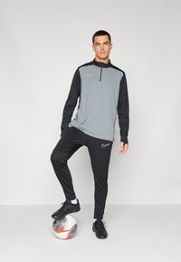 Grijze en zwarte sportieve trainingsoutfit met ritskraag, met Nike-logo's in oranje en wit. Model staat op een gemusterde voetbal.