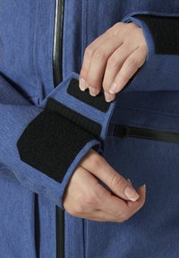 Blaue Jeansjacke mit schwarzen Klettverschlussmanschetten und einem Reißverschluss. Die Ärmel weisen ein strukturiertes Innenfutter und ein kantiges Manschettendesign auf.