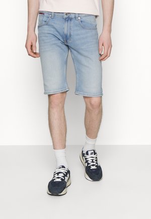 Homme portant un short en jean bleu clair, des chaussettes blanches et des baskets noires et blanches, debout devant un fond blanc.