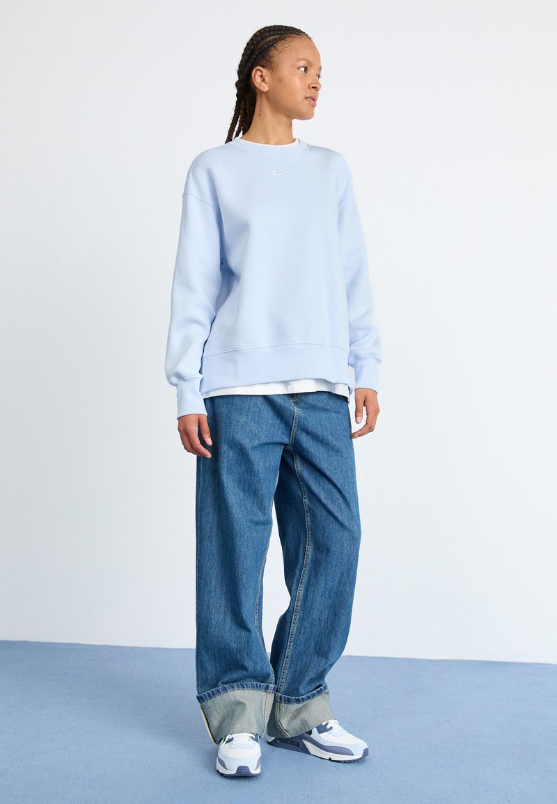 Sweatshirt azzurro chiaro con scollo rotondo e logo Nike bianco, abbinato a jeans in denim blu a gamba larga e scarpe da ginnastica bianche.