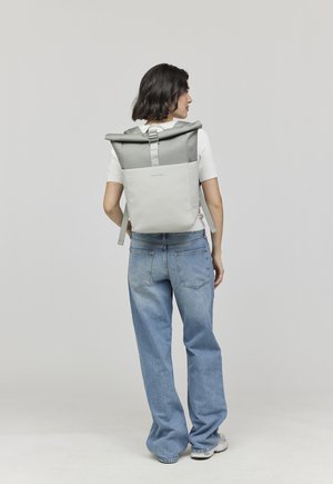 Femme portant un sac à dos à rabat gris clair et blanc, un t-shirt blanc à manches courtes, un jean bleu ample et des baskets blanches, se tenant devant un fond uni.