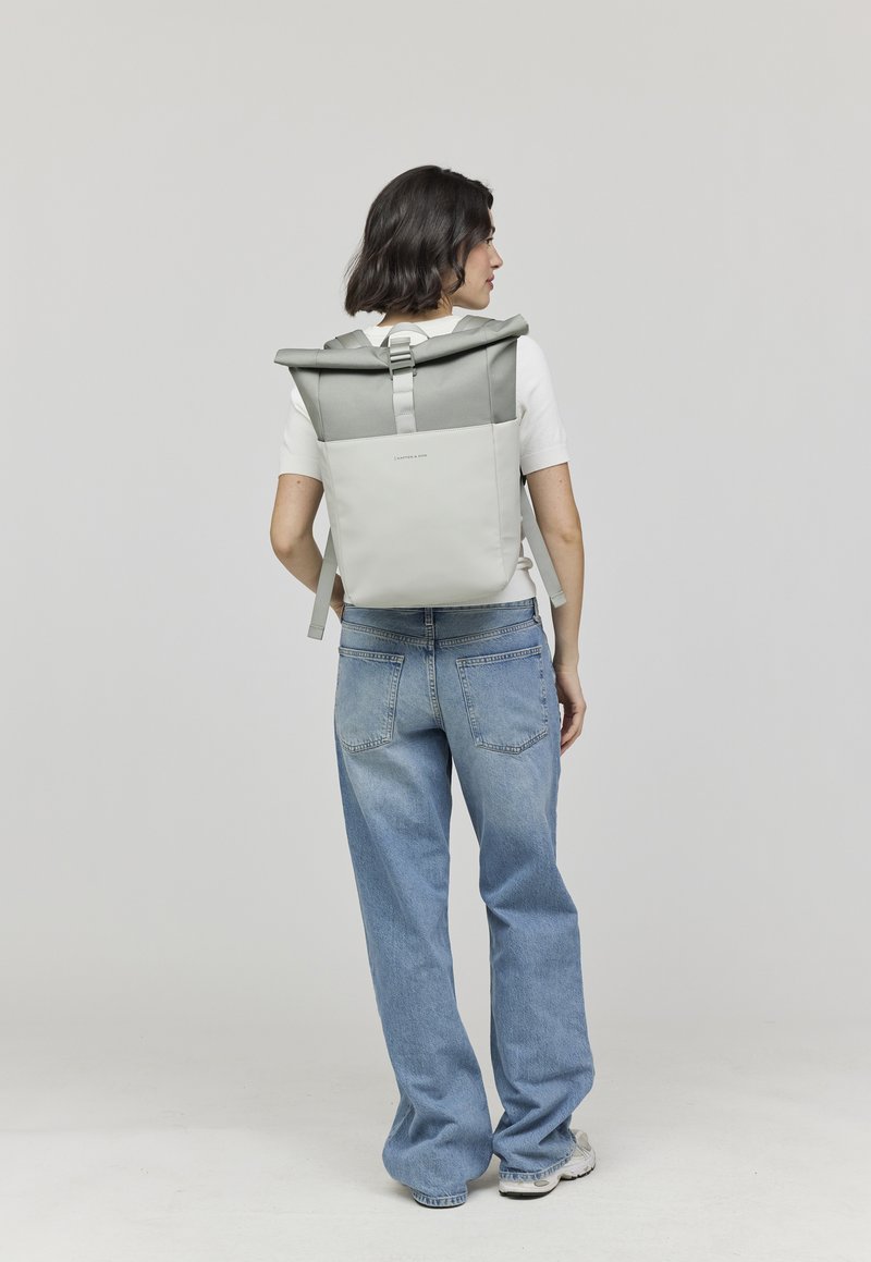 Femme portant un sac à dos à rabat gris clair et blanc, un t-shirt blanc à manches courtes, un jean bleu ample et des baskets blanches, se tenant devant un fond uni.