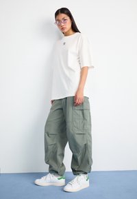 Junge Frau lehnt gegen eine weiße Wand und trägt ein übergroßes weißes Adidas-T-Shirt, lockere grüne Cargo-Hosen, weiße Sneakers und lila Cateye-Brille.