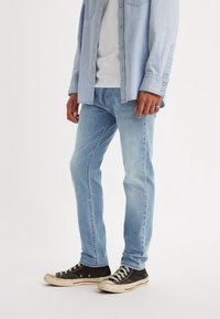 Jeans in denim blu chiaro con vestibilità dritta, abbinati a una camicia azzurro chiaro con bottoni e sneakers nere con suola in gomma bianca.