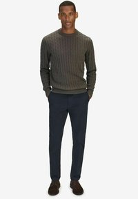 Donkergrijze kabelgebreide trui met ronde hals, gecombineerd met navy broek en brown suède schoenen, model staat met handen in de zakken.