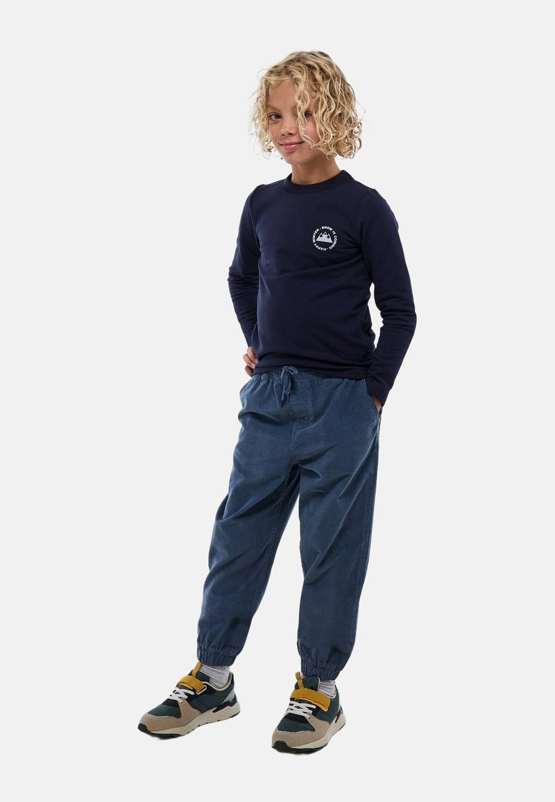Chemise à manches longues bleu marine avec un logo circulaire de montagne, associée à un pantalon bleu foncé ample avec ourlet élastique et des chaussures sportives.