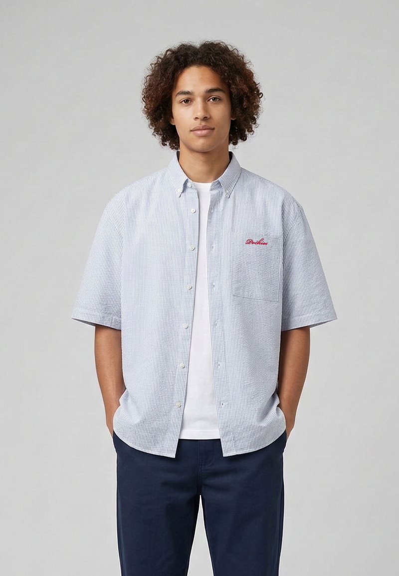 Jeune homme aux cheveux bouclés portant une chemise à manches courtes bleu clair par-dessus un t-shirt blanc, les mains dans les poches, debout devant un fond gris.