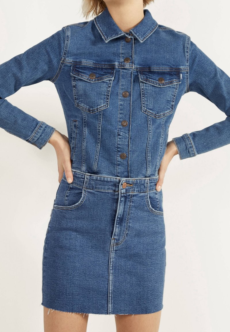 Kvinna som bär en blå denimjacka med knappar inskjutet i en matchande högmidjad denimkjol med fickor och fransad fåll.