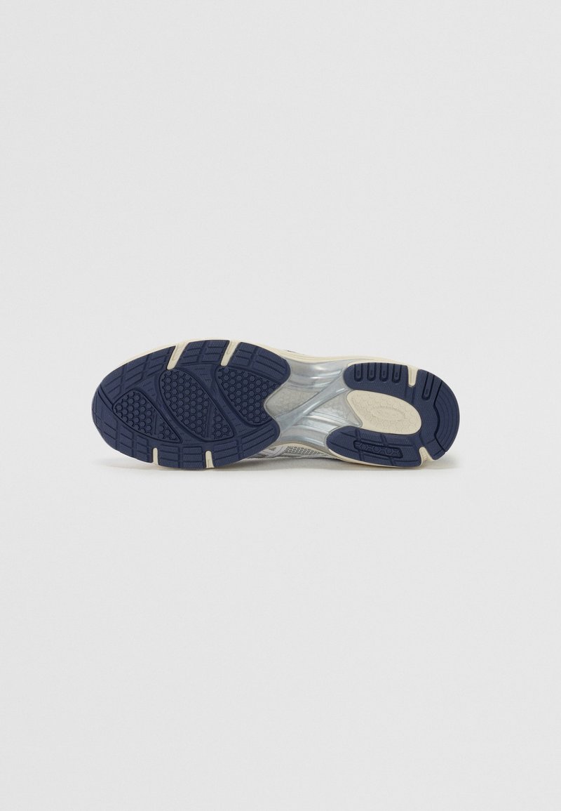 Suela de zapatilla con un patrón texturizado. Los colores incluyen azul marino y crema, con una sección transparente para amortiguación. Material de goma duradero.