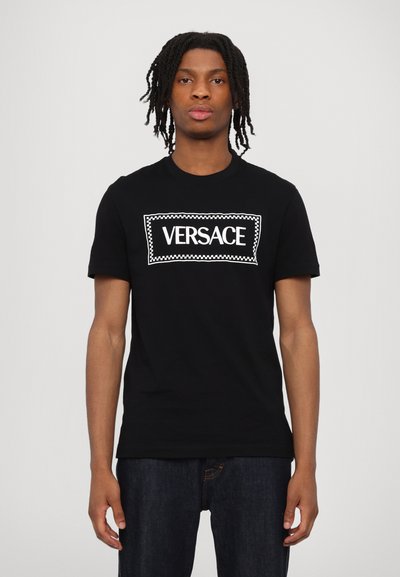 Musta puuvillainen t-paita, jossa on suuri valkoinen "VERSACE" -logo, joka on sijoitettu suorakulmaiseen kehykseen, jossa on ruutukuvio. Lyhyet hihat, pyöreä kaula-aukko.