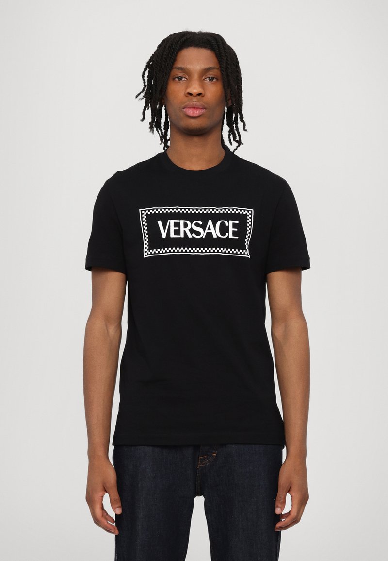 Melna kokvilnas T-kreklu ar lielu balto "VERSACE" logotipu, kas ir ietverts taisnstūrveida apmalē ar šaha rakstu. Īsās piedurknes, apaļa kakla izgriezums.