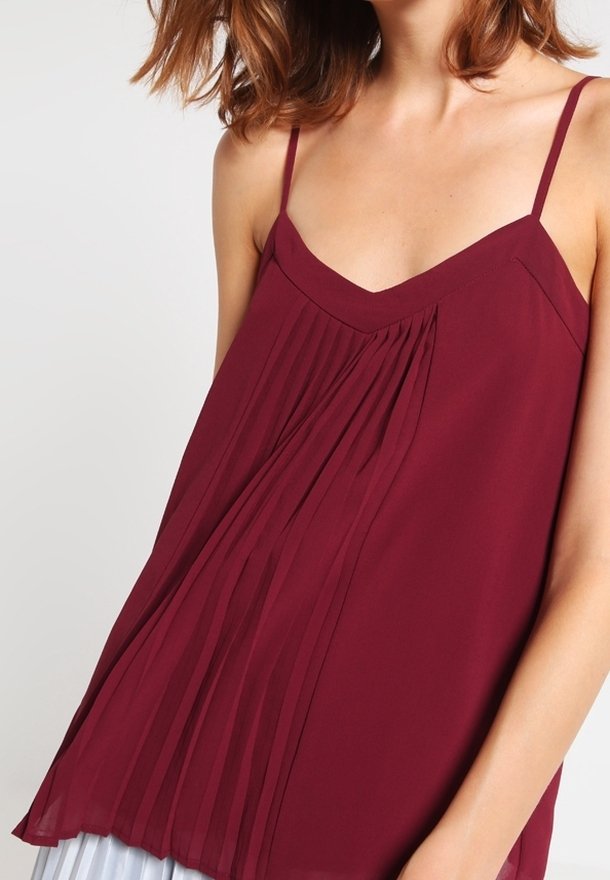 Blouse sans manches bordeaux avec un col en V, présentant des plis verticaux et des bretelles délicates, confectionnée dans un tissu léger et texturé.