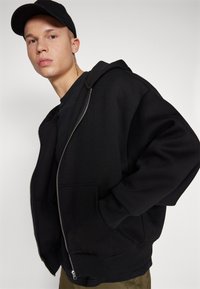 Black zip-up hoodie with a front pocket, featuring a loose fit, a drawstring hood, and ribbed cuffs. Worn over a black t-shirt.