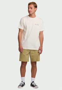 Jovem em pé voltado ligeiramente para a esquerda, vestido com uma t-shirt branca da Billabong, calções caqui, meias brancas e sapatilhas pretas.