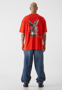 Rød bomulls t-skjorte med svart grafikk av en engel og tekst på baksiden. Kombinert med løse blå denimjogger og svarte sko.